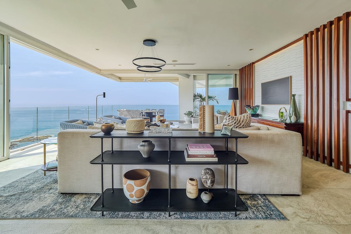 A modern living room with a large sofa, a coffee table, and a bookshelf. There is a view of the ocean through large windows.