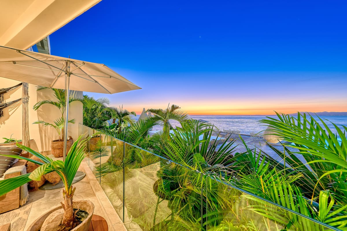 A balcony with a view of the ocean at sunset, surrounded by potted plants and a large umbrella.
