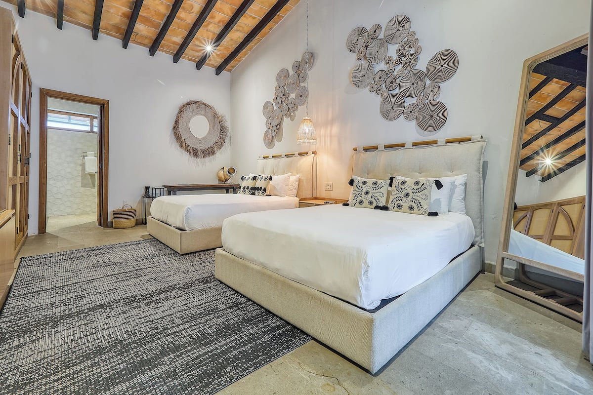 A bedroom with two beds, a wooden ceiling, a patterned rug, and wall art.