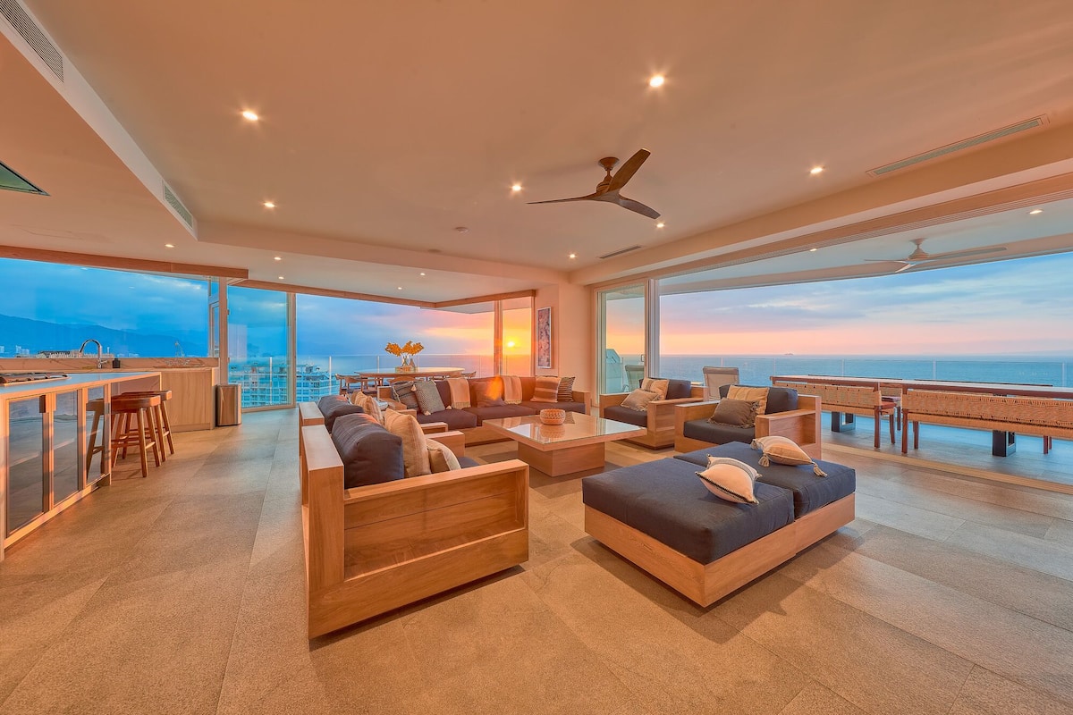 A spacious living area with a large window overlooking the sea, a ceiling fan, and a bar area.