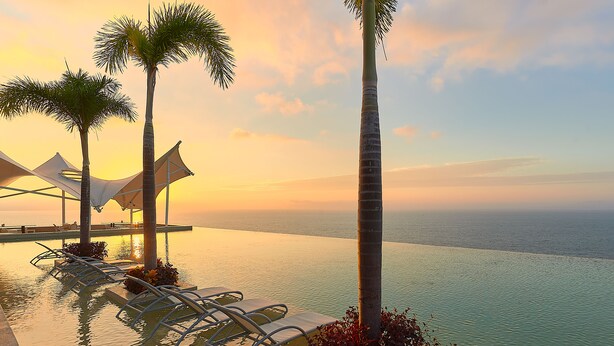 Piscina infinita con sillones de descanso y palmeras, con vistas al mar al atardecer.