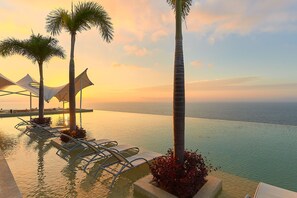 An infinity pool with lounge chairs and palm trees, overlooking the sea at sunset. - Maxwell Residences at Indah (Puerto Vallarta)