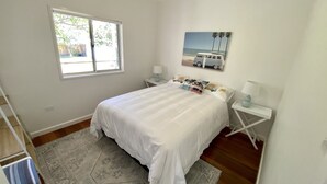 Ein Schlafzimmer mit Bett, Fenster, Nachttisch, Lampe und einem Bild mit Strandmotiv an der Wand.