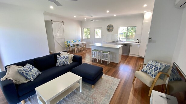 Ein modernes Wohnzimmer mit Sofa, Hocker und Couchtisch. Es gibt einen Essbereich mit Tisch und Stühlen sowie eine Küche mit Spüle und Arbeitsfläche.