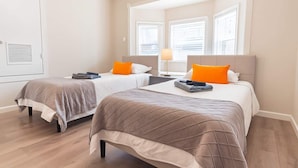 Une chambre d’hôtel avec deux lits, chacun doté d’une tête de lit et d’oreillers, une table de chevet avec une lampe, et une fenêtre avec des stores.