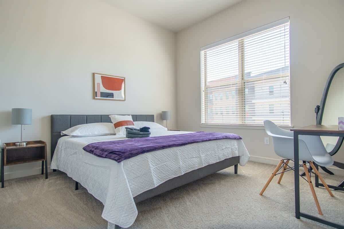 Une chambre à coucher comprenant un lit, des tables de chevet, un bureau, une chaise, une fenêtre avec des stores et une œuvre d’art accrochée au mur.
