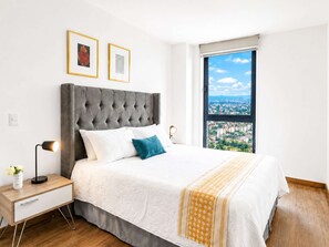 A bedroom with a large bed, a nightstand, a lamp, and a window with a city view.
