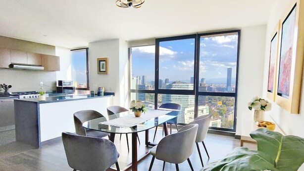Une cuisine moderne dotée d’un îlot central, d’un coin repas avec une table et des chaises, et de grandes fenêtres offrant une vue sur la ville.