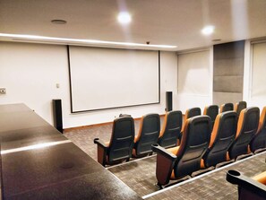 A conference room with a long table, rows of chairs, a projector screen, and speakers.