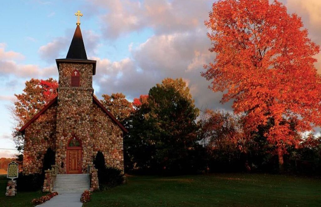 A stone church with a cross on top, surrounded by trees with autumn leaves.