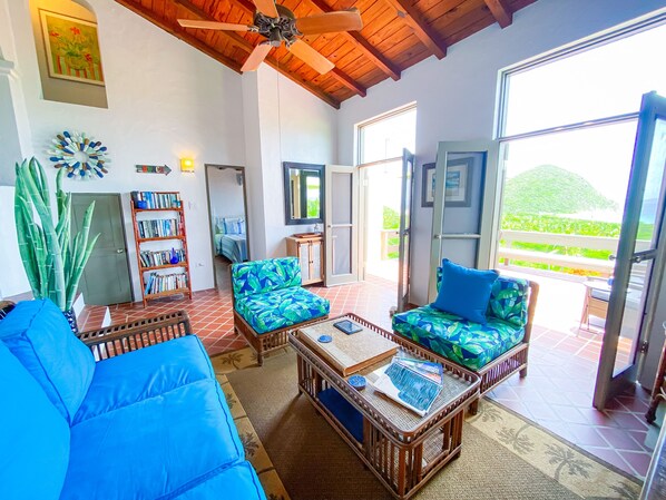 A spacious living area with a ceiling fan, a bookshelf, a large window with a view of greenery, and a door leading to a balcony. - Stunning panoramic views from the BVI (Tortola)