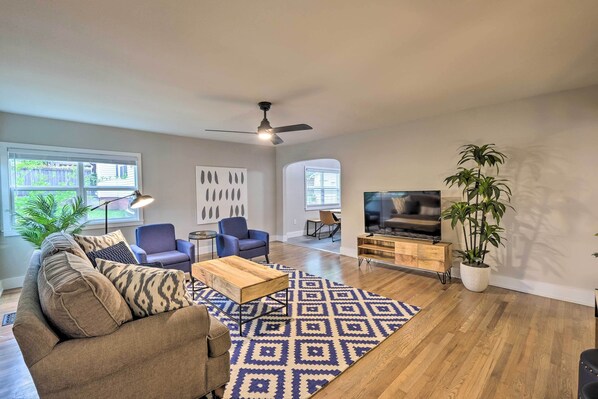 A spacious living room with a sofa, armchair, coffee table, and a patterned rug. There is a ceiling fan, a TV, and a potted plant. - Updated Fayetteville Home < 2 Miles to UArk! (Fayetteville)