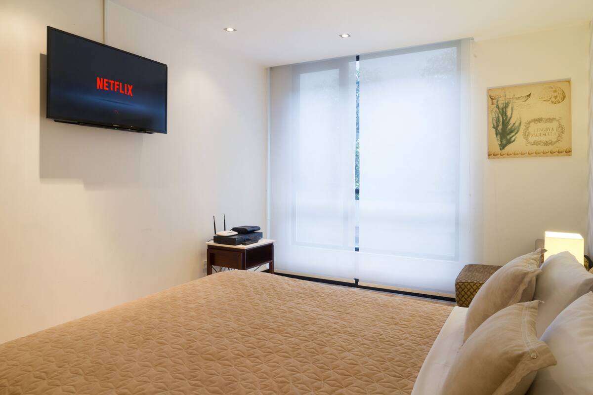 Quarto de hotel com uma cama, uma televisão exibindo 'NETFLIX', uma pequena mesa com um rádio e uma janela com persianas.