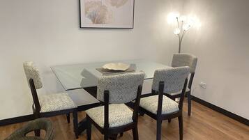 A dining area with a glass table surrounded by patterned chairs, a framed artwork above, and a vase with lights on the wall.
