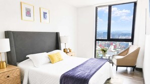 Een hotelkamer met een bed, een stoel, een nachtkastje, een lamp en een groot raam met uitzicht op de stad.