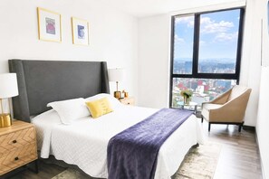Ein Hotelzimmer mit Bett, Stuhl, Nachttisch, Lampe und einem großen Fenster mit Blick auf die Stadt.