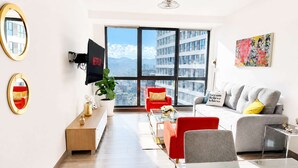 Een moderne woonkamer met een grote televisie, een bank, een rode fauteuil, een glazen salontafel en uitzicht op een stadsgezicht door grote ramen.