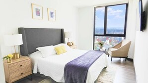 Hotellihuone, jossa on sänky, yöpöydät, tuoli, ikkuna, josta on näkymä kaupunkiin, sekä seinällä olevia kehystettyjä tauluja.