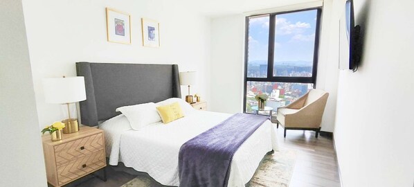 Ein Hotelzimmer mit Bett, Nachttischen, einem Sessel, einem Fenster mit Blick auf die Stadt und gerahmten Bildern an der Wand.