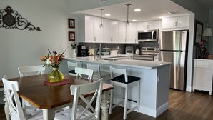 A modern kitchen with white cabinetry, a central island, stainless steel appliances, and a dining area with white chairs and a wooden table. - Lovely renovated condo with water views, great value for outstanding amenities (Clearwater Beach)