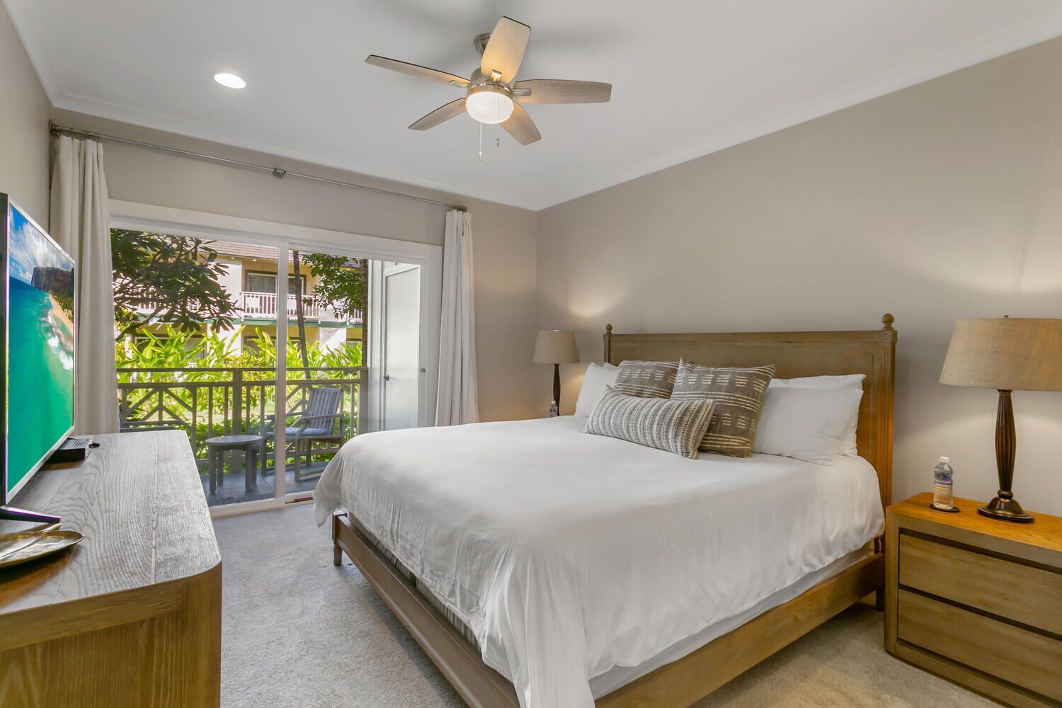 A hotel room with a bed, a desk, a TV, a ceiling fan, and a balcony with a view of greenery.