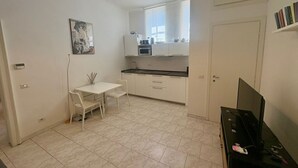 Una habitación pequeña y limpia con escritorio blanco, sillas, televisor y kitchenette.