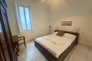 Un dormitorio con cama, una silla, un ventanal y una mesita de noche.