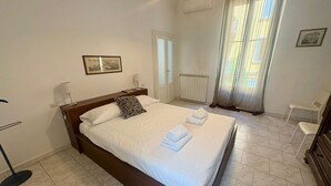 Una cama bien hecha con almohadas y toallas dobladas, una mesita de noche con lámpara, un cuadro en la pared, un radiador y una ventana con cortinas.