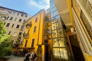 Une rue étroite en milieu urbain, avec un immeuble jaune doté de balcons et d’une façade en verre.