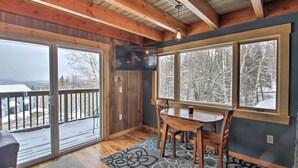 Una habitación acogedora con techo de madera, un televisor grande montado en la pared, una mesa de comedor con sillas y vistas a árboles nevados a través de amplios ventanales.