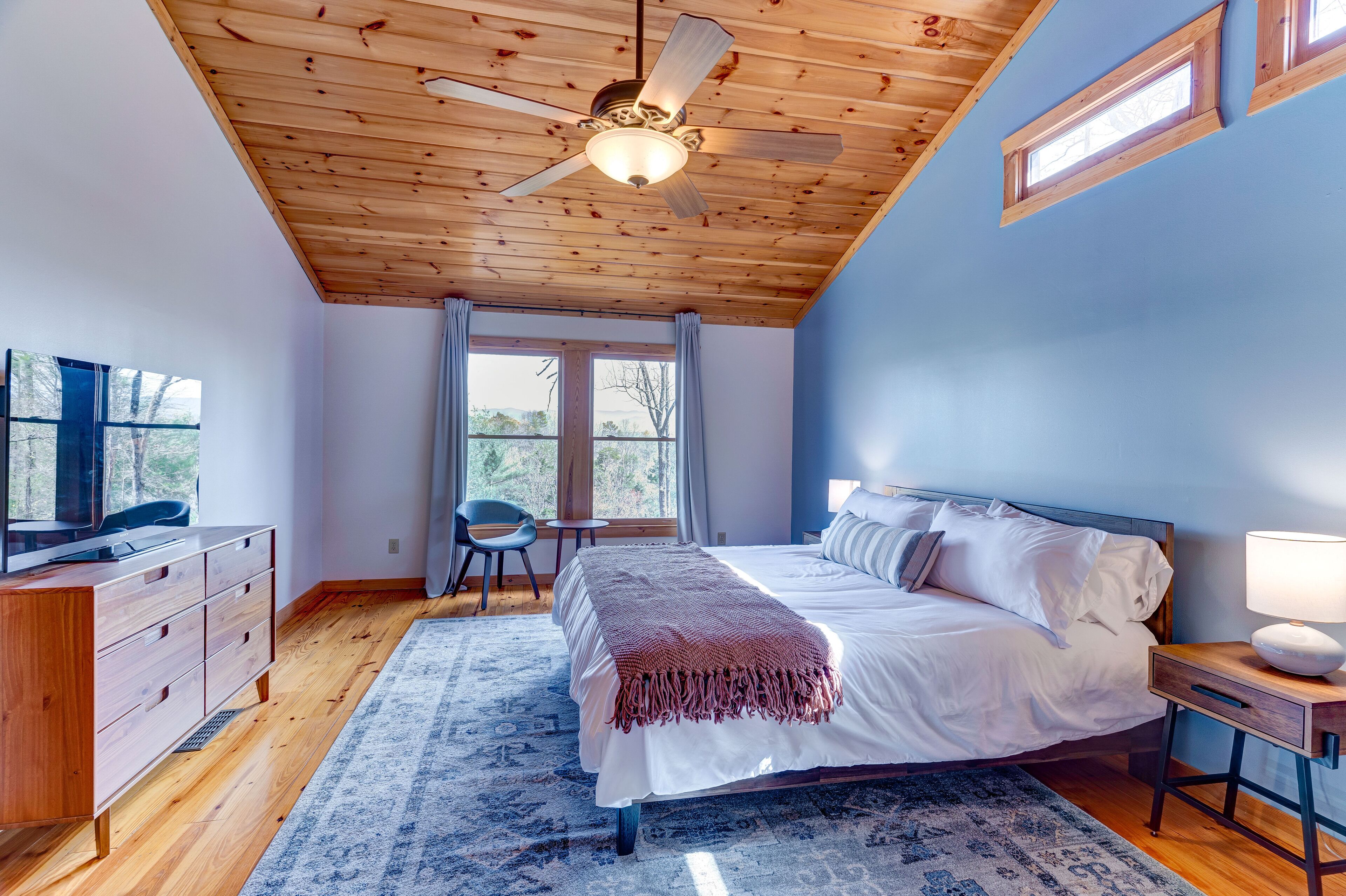 A bedroom with a wooden ceiling, a bed with white bedding, a nightstand with a lamp, a TV on a wooden dresser, and a view of the outdoors through a window.