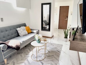 Una sala de estar moderna con un sofá gris, una mesita redonda con un florero y una alfombra de diseño en patrón de espiga, además de un espejo grande en la pared.