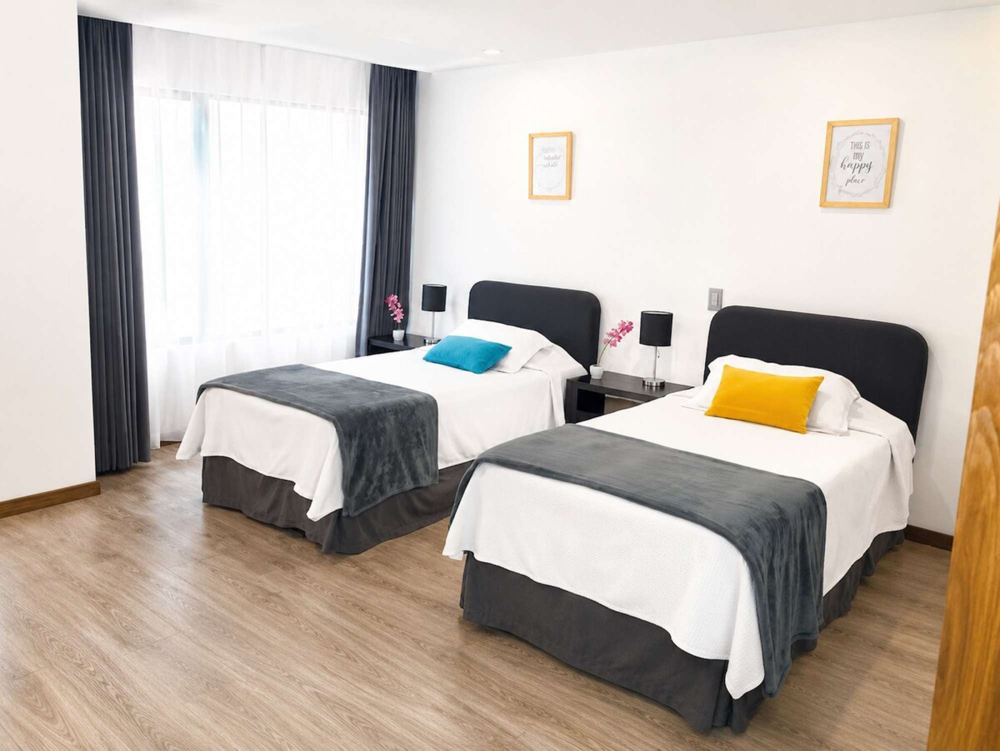 A hotel room with two beds, a window with curtains, and a wall with two framed pictures.