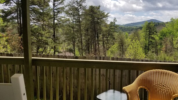 Una terraza de madera con una silla de mimbre y una mesa de vidrio, con vistas a una zona boscosa y montañas a lo lejos.