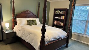 Una cama con dosel y tablero de madera, una mesita de noche con lámpara, un cojín decorativo verde y una estantería llena de libros.