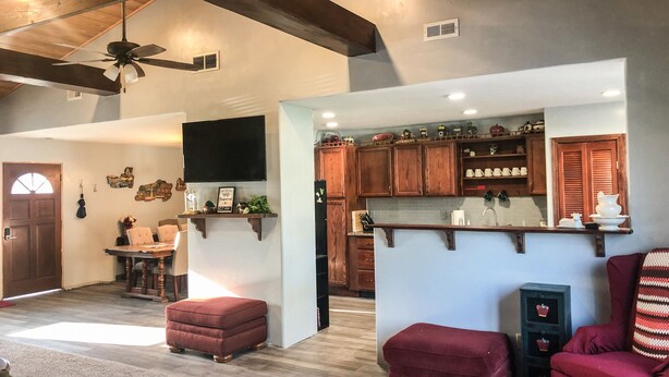 A spacious living area with a wooden ceiling, a flat-screen TV mounted on the wall, and a kitchen with wooden cabinets and a countertop.