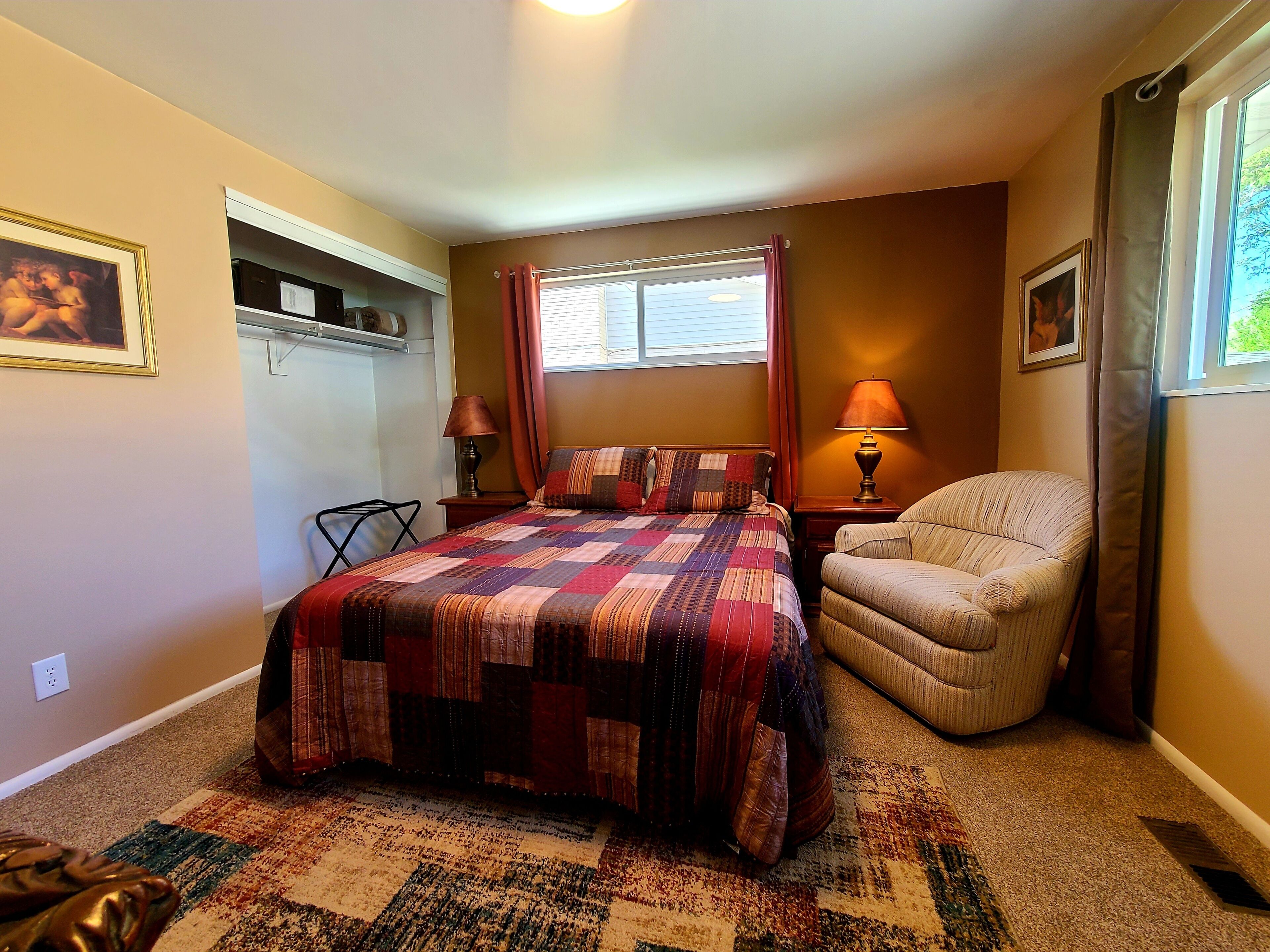 Un dormitorio con cama, una silla, ventana con cortinas, lámpara y estante con objetos.