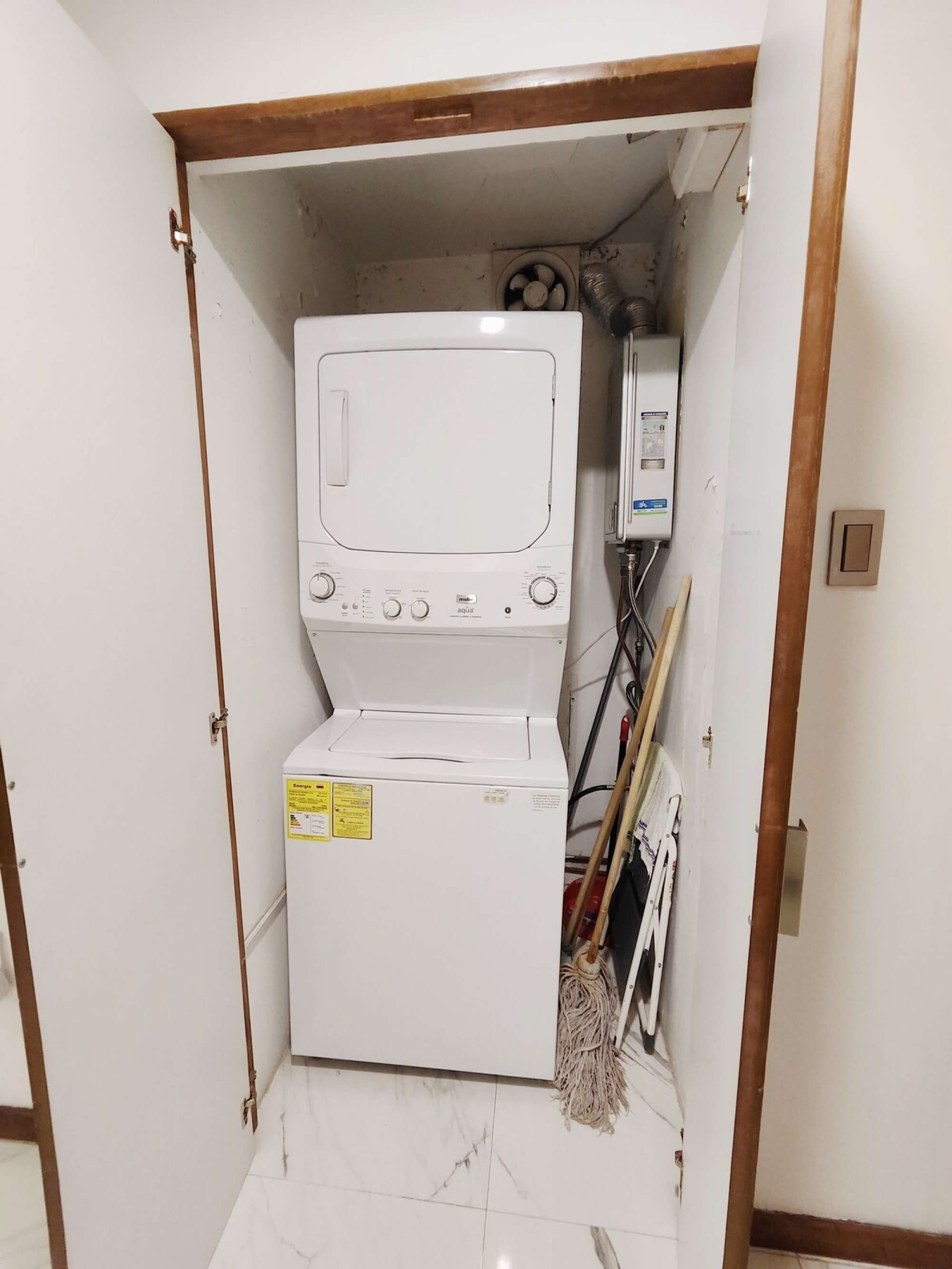 A compact laundry room with a front-loading washer and dryer, a utility meter, and a mop leaning against the wall.