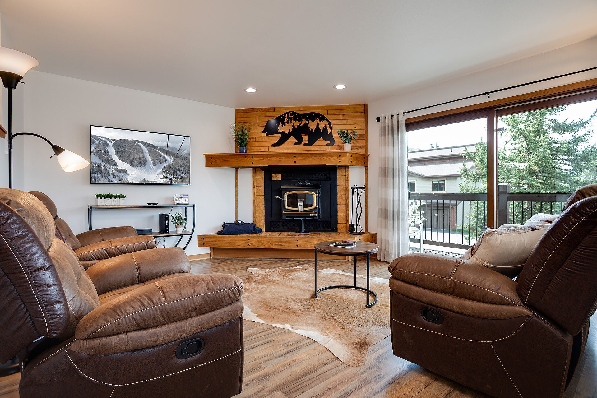 Una sala de estar con sofás de cuero, chimenea, televisor montado y un ventanal con vistas a árboles.