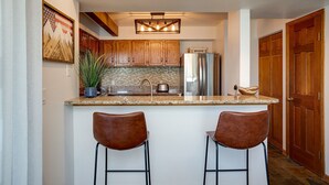 Una cocina con armarios de madera, encimera de granito, refrigerador de acero inoxidable, lámpara colgante y dos taburetes de bar.