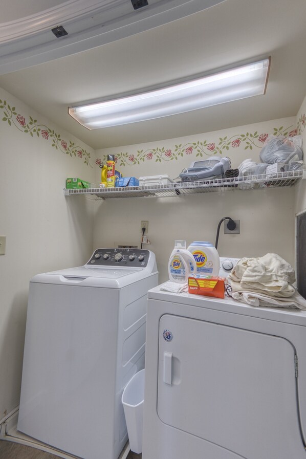 Un espace buanderie comprenant un lave-linge, un sèche-linge, des étagères avec des produits de lessive et une poubelle.