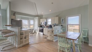 A spacious kitchen with white cabinets, a dining area with a wooden table and chairs, and a view of the outdoors through large windows. - Captain's Quarters (Frederica)