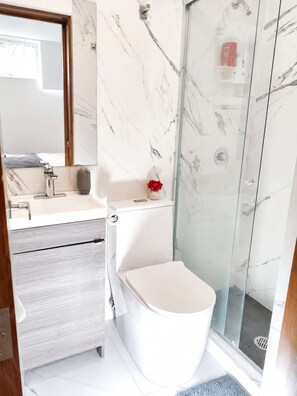 Ein Badezimmer mit Marmorwand, Toilette, Waschbecken inklusive Spiegel und einer Duschkabine aus Glas.