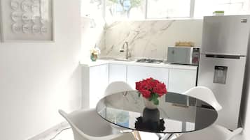 Cozinha moderna com bancada branca, fogão a gás, micro-ondas e geladeira. Há uma mesa de jantar com duas cadeiras e um vaso com flores vermelhas.
