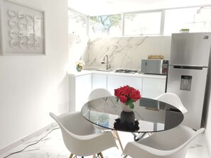 Eine moderne Küche mit weißer Arbeitsplatte, einem Gasherd, einer Mikrowelle und einem Kühlschrank. Es gibt einen Esstisch mit zwei Stühlen und eine Vase mit roten Blumen.