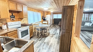 A wooden cabin interior with a kitchen, dining area, and a view through a door.