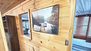 A cabin interior with wooden walls, a mounted TV, and a view of a lake through a window.