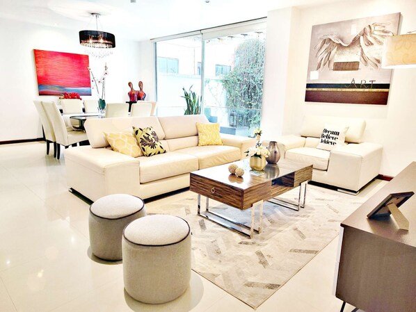 Un salon moderne avec un canapé beige, une table basse et deux poufs. Un tableau orne le mur, et une fenêtre offre une vue sur la verdure.
