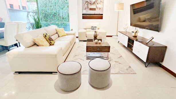 Un salon moderne avec un canapé blanc, une table basse et deux poufs. Un téléviseur à écran plat est fixé au mur, et un tableau est accroché au-dessus du canapé.