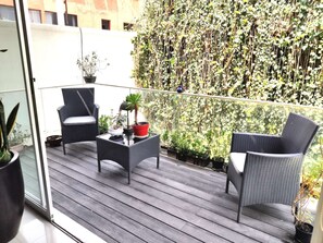 Een balkon met rieten meubels, planten in potten en een weelderige groene haag op de achtergrond.